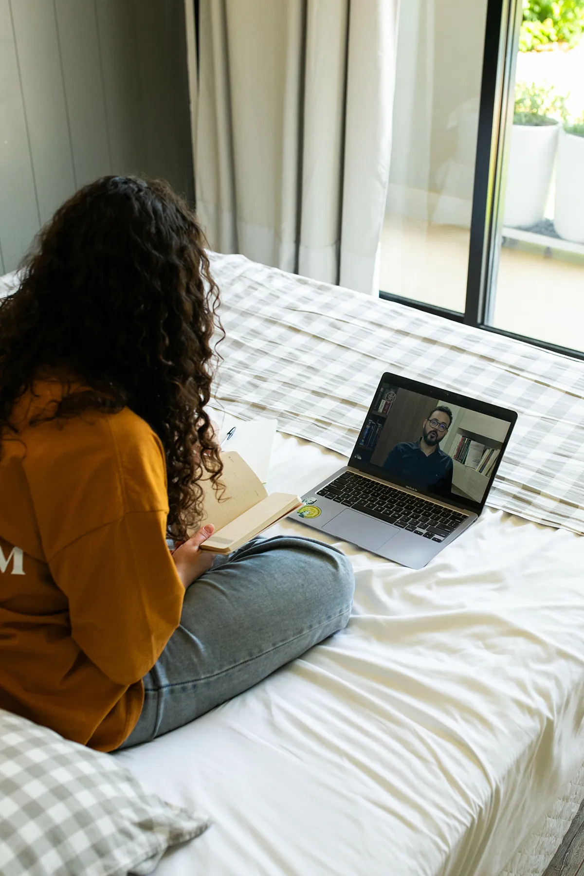 Mujer sentada en la cama, con un cuaderno sobre las piernas, viendo una clase en la computadora portátil